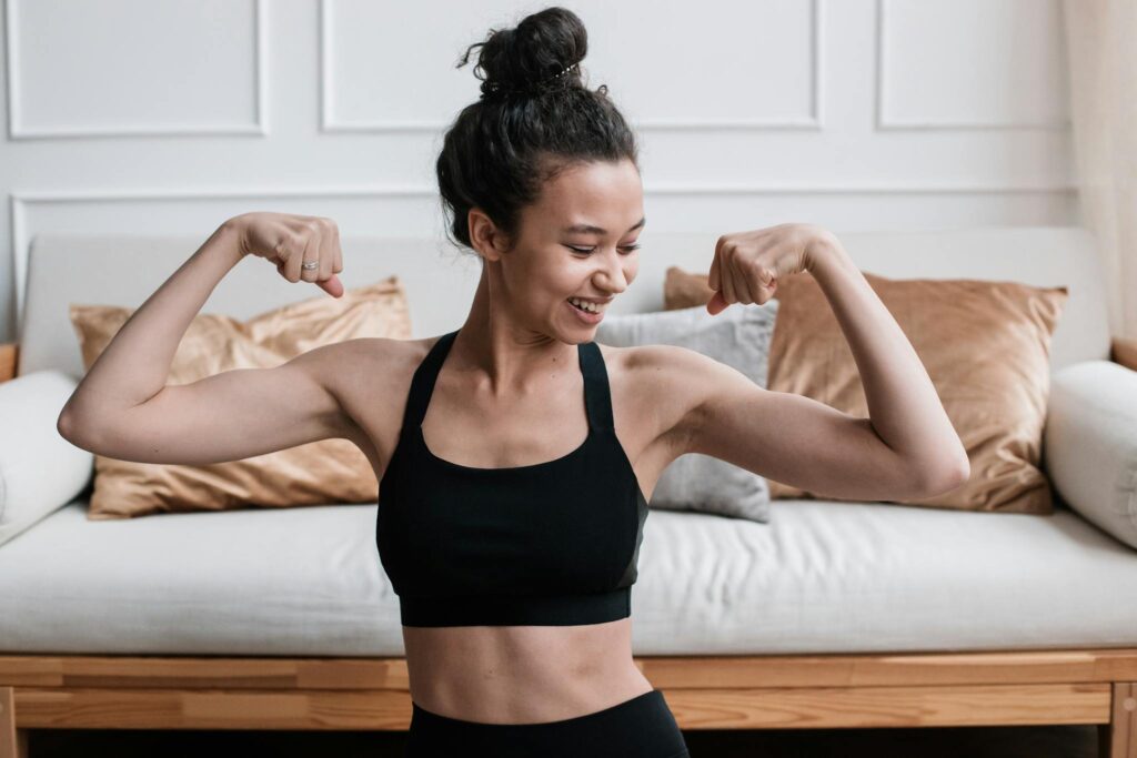 Smiling woman flexing her muscles at home in a sporty outfit, showcasing fitness and strength.
